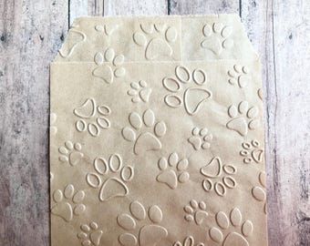 May include: A light brown paper bag featuring an embossed pattern of various sized animal paw prints across the entire surface. The bag has a flat, rectangular body with a folded top edge, showcasing a repeating texture of raised paw prints.