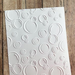 May include: White cardstock with an embossed bubble pattern. The design features various sizes of overlapping circles, creating a textured, bubbly effect. The paper is set against a wooden background.