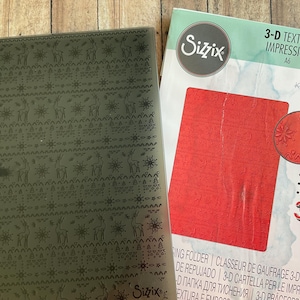 May include: A gray plastic Sizzix 3-D Textured Impressions A6 embossing folder featuring a repeating pattern of deer and snowflakes, accompanied by its original white and light green retail packaging displaying a red sample of the embossed design.