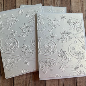 May include: A stack of white greeting cards with an embossed snowflake design. The cards feature a repeating pattern of snowflakes and swirling lines, creating a wintery theme. The cards are on a wooden surface.