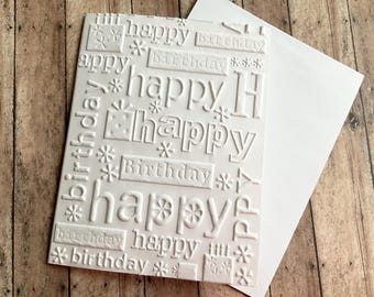 May include: White embossed birthday card with the words "happy" and "birthday" in various fonts and sizes. The card is accompanied by a white envelope, set against a wooden background.