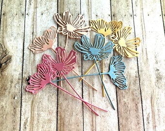 May include: A set of seven decorative metal flowers in various colors, including pink, blue, and yellow. The flowers are attached to thin metal stems and have a delicate, intricate design.