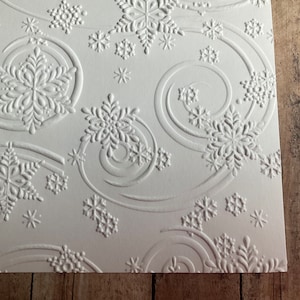 May include: White embossed paper with a repeating snowflake pattern. The design features swirling lines and various sizes of snowflakes, creating a textured, winter-themed aesthetic. The paper is a bright white color.