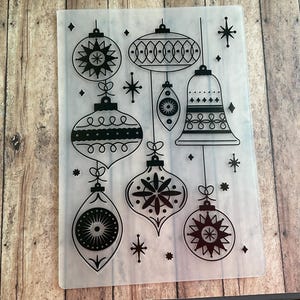 May include: A rectangular clear plastic embossing folder featuring a black printed design of various hanging Christmas ornaments including baubles, a bell, and starburst accents. The pattern is arranged vertically with multiple shapes.