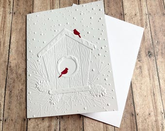 May include: A white greeting card featuring an embossed design of a wooden birdhouse surrounded by pine needles and pinecones. Two small, bright red cardinal birds are positioned on the birdhouse, with embossed snow dots throughout.