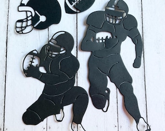 May include: Black paper cutouts of football-themed elements. Includes a football helmet, a football, and two football players in action poses. The cutouts are arranged on a light wood-grain surface.