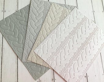 May include: Four textured paper sheets in shades of gray, beige, and white. Each sheet features a raised cable knit pattern. The sheets are arranged diagonally, showcasing the different color variations and the detailed design.