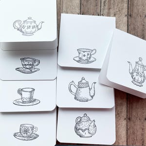Puede incluir: Un conjunto de tarjetas blancas con ilustraciones en tinta negra de tazas de té y teteras. Las tarjetas están apiladas y desplegadas, mostrando los diferentes diseños. El fondo es una superficie de madera.
