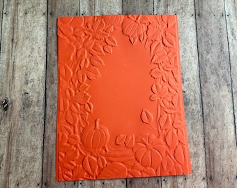 May include: A rectangular orange embossing folder featuring a raised, textured design of pumpkins and autumn leaves arranged in a border around a smooth, blank center space, creating a relief pattern for crafting and paper projects.