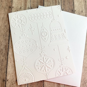 May include: A white embossed Christmas card featuring various ornament designs, including bells and starbursts. The card is paired with a blank white envelope, perfect for holiday greetings. The card is on a wooden surface.