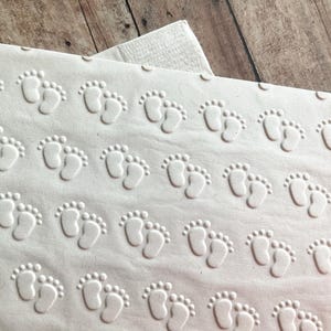 Puede incluir: Servilletas de papel blancas con un patrón en relieve de pequeñas huellas de pies. Las servilletas están dobladas y apiladas.