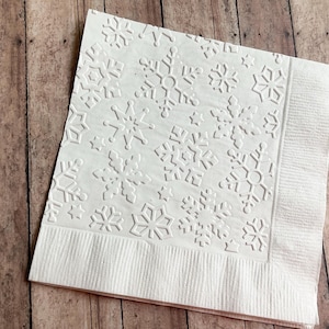 May include: White paper napkin with an embossed snowflake pattern. The napkin has a textured border and is set against a wooden background. Ideal for winter or holiday events.