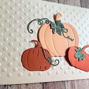 May include: A decorative card featuring three paper pumpkins in shades of orange and peach, with green vines and leaves. The background is a light beige with a dotted pattern, creating a fall-themed design.