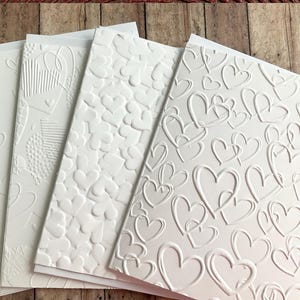 May include: A stack of white greeting cards with embossed heart designs. The cards feature various heart patterns, including scattered hearts, textured hearts, and overlapping hearts. The cards are arranged on a wooden surface.