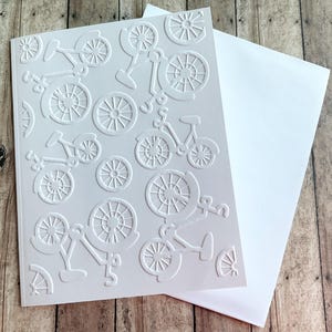 May include: A white greeting card with an embossed bicycle design, accompanied by a white envelope. The card showcases a repeating pattern of bicycles and wheels. The card and envelope are on a wooden surface.