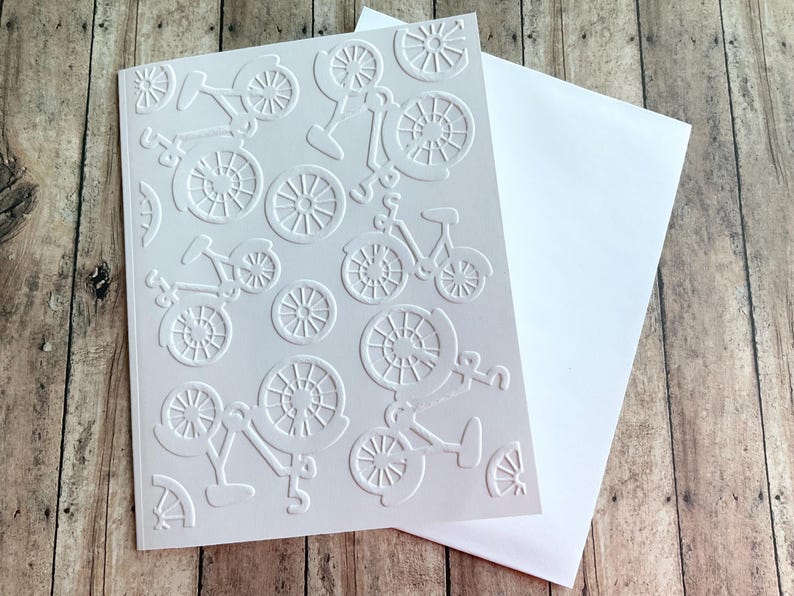 May include: A white greeting card with an embossed bicycle design, accompanied by a white envelope. The card showcases a repeating pattern of bicycles and wheels. The card and envelope are on a wooden surface.