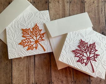May include: Two white cards with a fall leaf design. The cards have a raised leaf pattern and a single, larger leaf in the center. One card has an orange leaf and the other has a burgundy leaf.