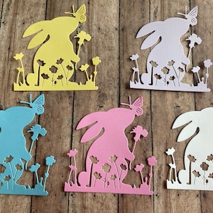 May include: Five paper cutouts of bunnies in different colors: yellow, lavender, light blue, pink, and white. Each bunny is holding a flower and has a butterfly on its nose. The bunnies are all in a field of flowers.