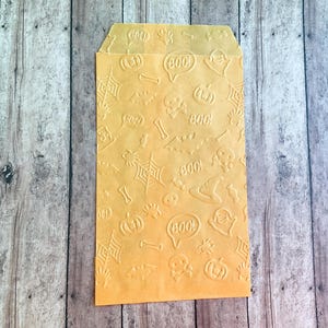 Puede incluir: Una bolsa de papel rectangular de color naranja con un patrón en relieve de iconos temáticos de Halloween, que incluyen calabazas, telarañas, murciélagos, calaveras, huesos, sombreros de bruja y globos de texto con la palabra BOO.