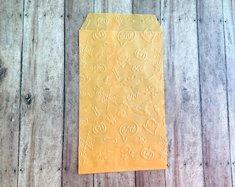 May include: A rectangular orange paper bag featuring an embossed pattern of Halloween-themed icons including pumpkins, spider webs, bats, skulls, bones, witch hats, and speech bubbles containing the text BOO. The bag has a flat top edge.