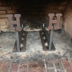 May include: Two metal fireplace log holders with the letter "H" on each. The holders are made of black metal and have a rustic finish. They are sitting in front of a brick fireplace.