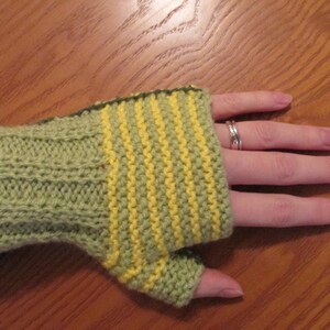 Könnte beinhalten: Handgestrickte Fingerhandschuhe in Grün- und Gelbtönen. Der Handschuh hat eine gerippte Manschette und vertikale gelbe Streifen auf der Handfläche. Das offene Fingerdesign ermöglicht Geschicklichkeit und hält die Hände warm. Ein gemütliches Accessoire für kühleres Wetter.