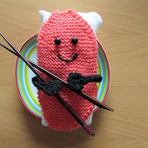 Könnte beinhalten: Ein rosa gestricktes Sushi mit einem lächelnden Gesicht, schwarzen Augen und einem schwarzen Schnurrbart. Das Sushi liegt auf einem bunten Teller mit zwei Holzstäbchen.
