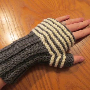 Könnte beinhalten: Fingerlose Handschuhe in Grau und Weiß mit einem grauen Bündchen. Die Handschuhe sind gestrickt und haben ein strukturiertes Muster.