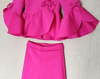 May include: A bright pink two-piece outfit for girls. The top has a ruffled neckline and long sleeves with a bow detail on each sleeve. The pants have a ruffled hem.