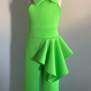Op de afbeelding: Een fel limoengroene jumpsuit met een halternek en een uitlopende kraag. De jumpsuit heeft een getailleerd lijfje, een brede tailleband en een gefranjerde accent aan de zijkant. De broek heeft een uitlopend pijp ontwerp.