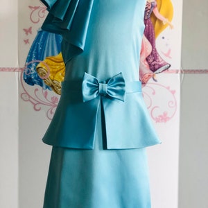 May include: A light blue dress with a bow and layered skirt. The sleeveless dress features a ruffled shoulder detail and a bow at the waist. The dress is made of a smooth, satin-like fabric. The dress is displayed on a mannequin.