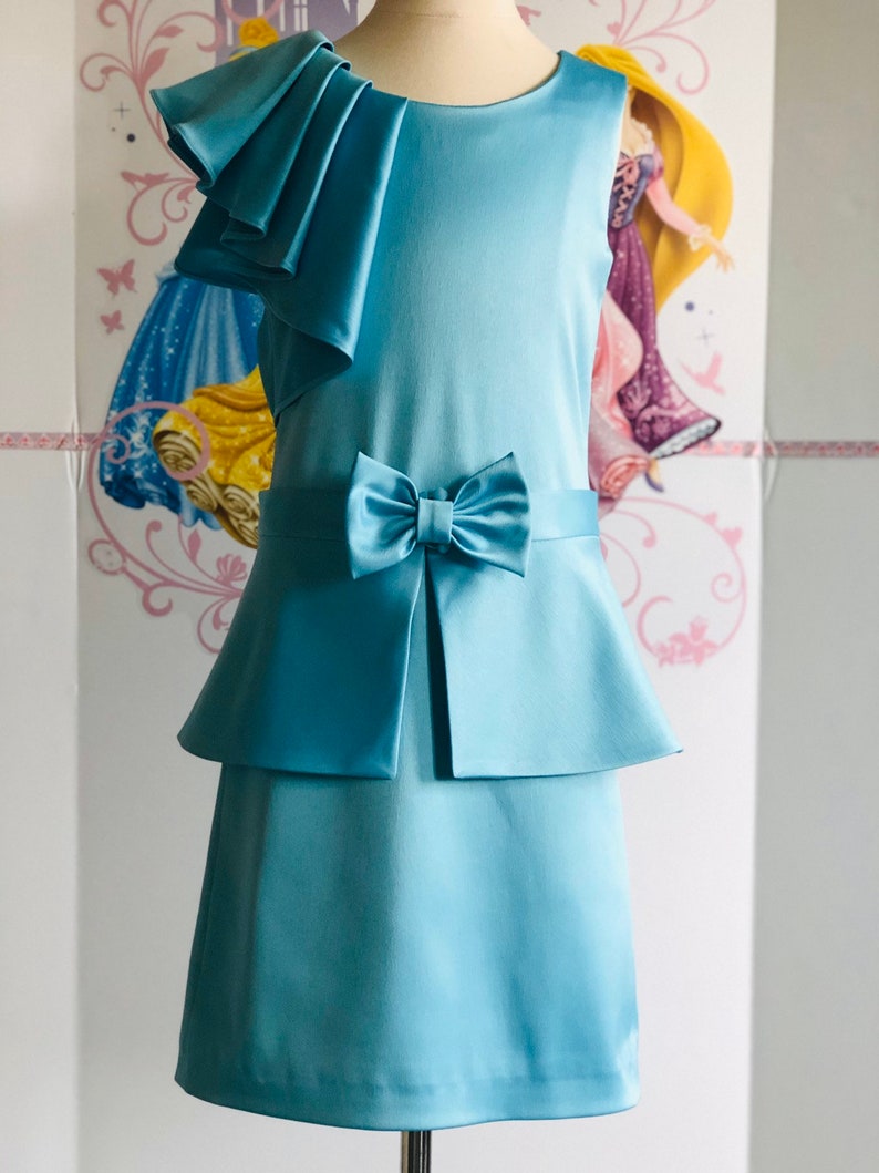 May include: A light blue dress with a bow and layered skirt. The sleeveless dress features a ruffled shoulder detail and a bow at the waist. The dress is made of a smooth, satin-like fabric. The dress is displayed on a mannequin.