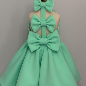 Op de afbeelding: Een mintgroene jurk met drie grote strikken op de rug. De jurk heeft een volle rok en is gemaakt van een zachte, vloeiende stof.