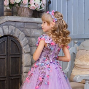 May include: A young girl wearing a lavender tulle dress with floral embroidery and a flower crown. The dress has pink, blue, and lavender flowers. The girl is standing in front of a stone wall with a wooden door.