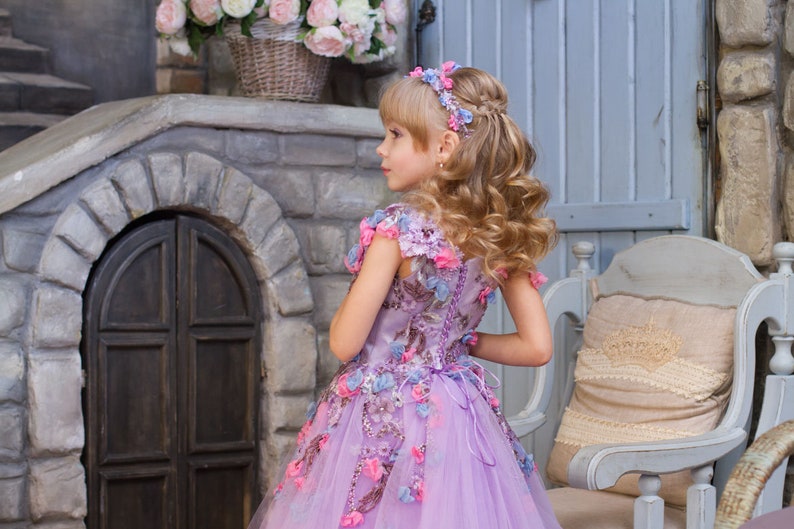 May include: A young girl wearing a lavender tulle dress with floral embroidery and a flower crown. The dress has pink, blue, and lavender flowers. The girl is standing in front of a stone wall with a wooden door.