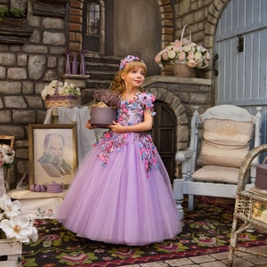 May include: A young girl in a lavender tulle dress with floral embellishments holds a decorative box. She is standing in a room with stone walls, a fireplace, and a wicker chair.