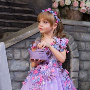 May include: A young girl in a lavender dress with floral embroidery holds a purple teapot. The dress has a tulle skirt and is decorated with pink, blue, and lavender flowers. The girl is wearing a flower crown.