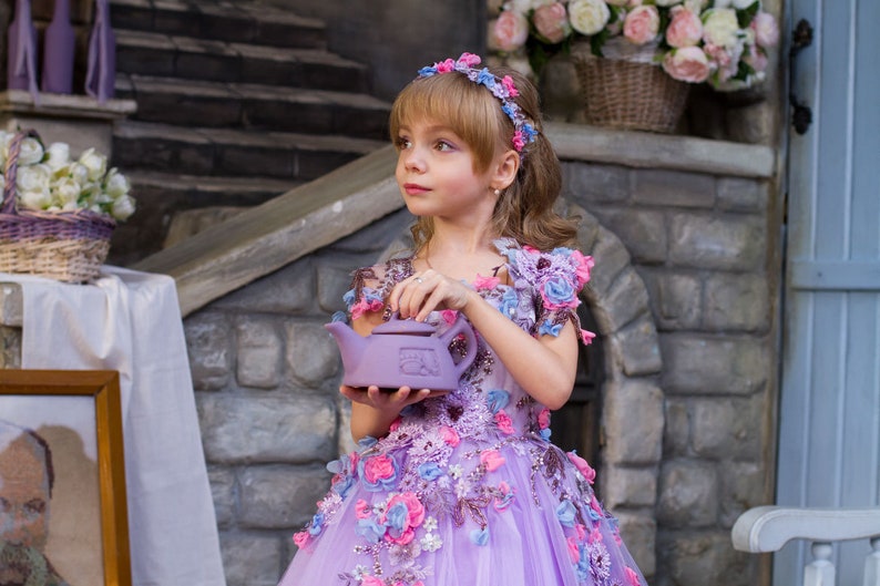 May include: A young girl in a lavender dress with floral embroidery holds a purple teapot. The dress has a tulle skirt and is decorated with pink, blue, and lavender flowers. The girl is wearing a flower crown.