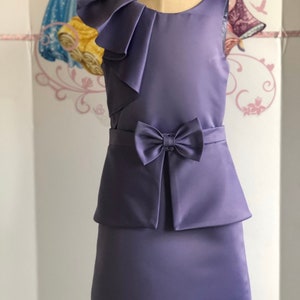 May include: A lavender sleeveless dress with a bow at the waist and a layered skirt. The dress features a ruffled shoulder detail. The dress is displayed on a mannequin. The background has a princess theme.