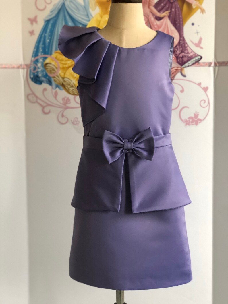 May include: A lavender sleeveless dress with a bow at the waist and a layered skirt. The dress features a ruffled shoulder detail. The dress is displayed on a mannequin. The background has a princess theme.
