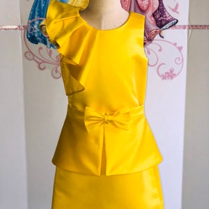 May include: A bright yellow dress with a layered skirt and a bow at the waist. The sleeveless top features a ruffled shoulder detail. The dress is displayed on a mannequin, showcasing its design and fit.