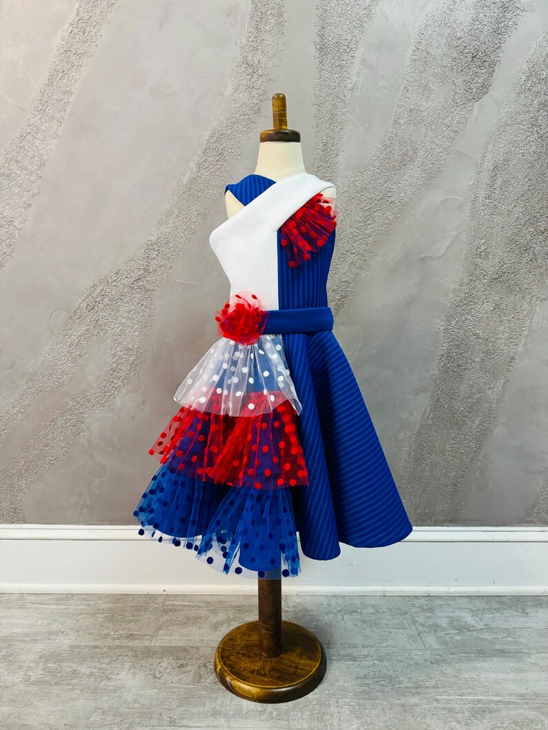 May include: A blue and white dress with red and white polka dot tulle layers. The dress has a white bodice with a blue stripe and a red and white tulle flower.