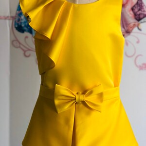 May include: A bright yellow dress with a bow and ruffle detail. The sleeveless dress features a fitted bodice, a bow at the waist, and a layered skirt. The dress is made of a smooth, satin-like fabric and is displayed on a mannequin.