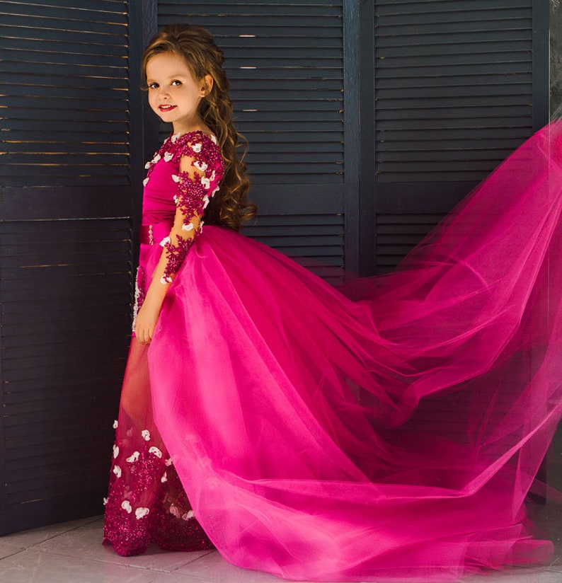 May include: A young girl wearing a long, pink tulle dress with a fitted bodice and lace sleeves. The dress has a train and is decorated with floral embellishments.
