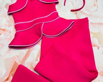 May include: A pink outfit with a halter top, pants, a headband, and a bow. The top has a ruffled neckline and a pink trim. The pants are wide-legged and have a pink trim. The headband is pink and the bow is pink with a white center.
