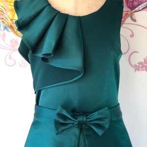May include: A sleeveless, emerald green dress with a satin finish. The dress features a ruffled accent on the shoulder and a bow detail at the waist. The skirt has a slight A-line shape.