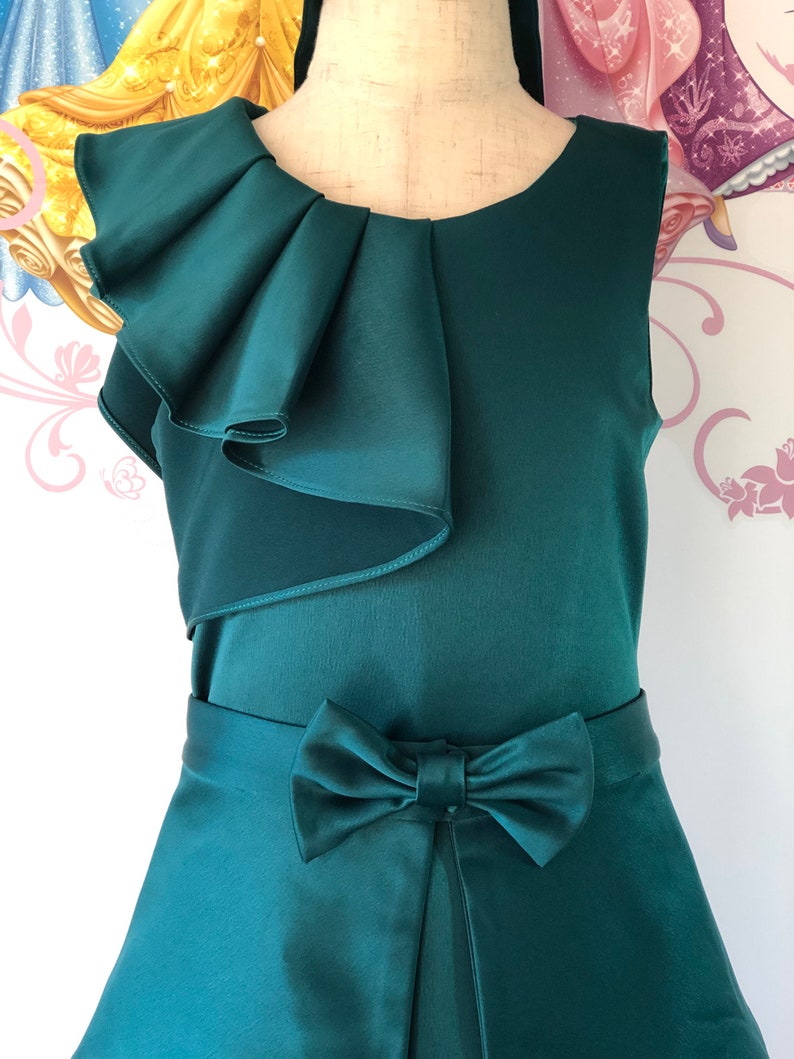 May include: A sleeveless, emerald green dress with a satin finish. The dress features a ruffled accent on the shoulder and a bow detail at the waist. The skirt has a slight A-line shape.