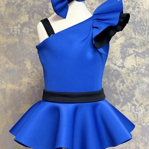Puede incluir: Un vestido de baile azul y negro de un solo hombro con una manga con volantes y un cinturón negro. El vestido tiene una falda completa y se lleva en un maniquí con un lazo azul.