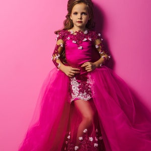 May include: A young girl wearing a pink dress with a white lace overlay and a tulle skirt. The dress is decorated with pink and white flowers. The girl is standing in front of a pink background.