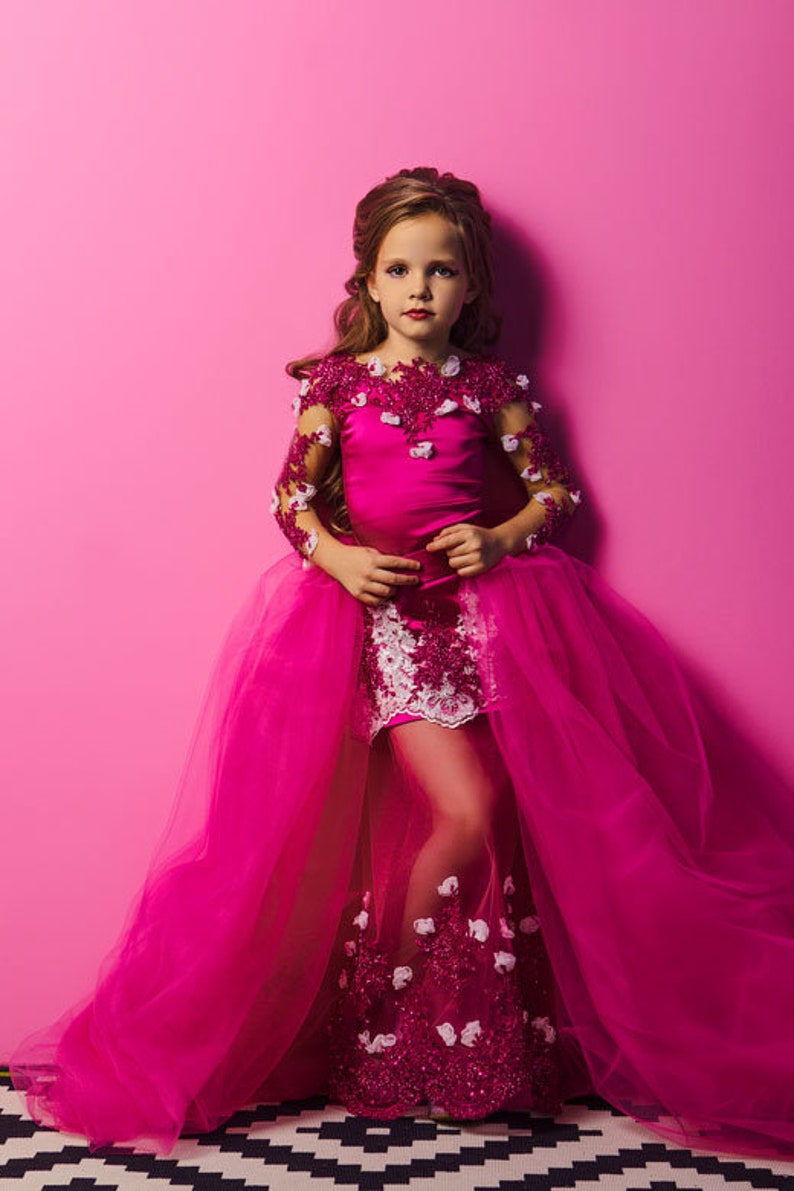 May include: A young girl wearing a pink dress with a white lace overlay and a tulle skirt. The dress is decorated with pink and white flowers. The girl is standing in front of a pink background.